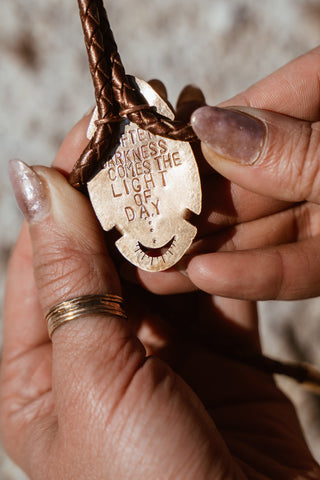 Hand holding a brass pendant with engraved text against a blurred background